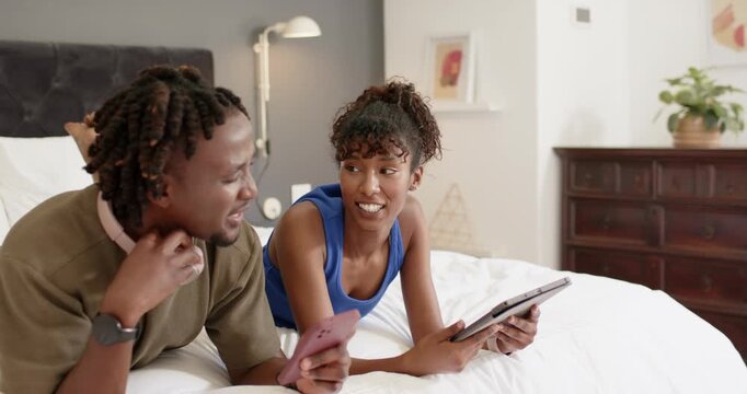 African American couple lying in bed woman showing gray tablet nudging man with pink phone leaning