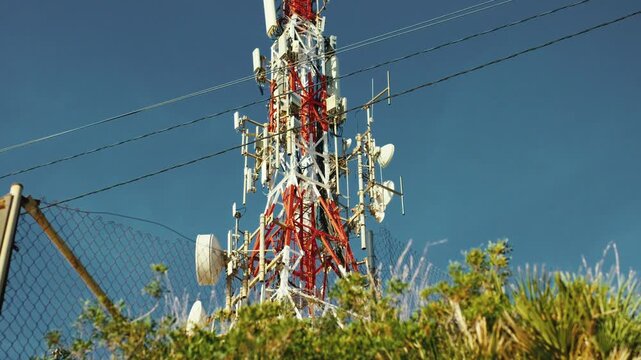 Telecommunications tower against clear blue sky with surrounding foliage and fence in foreground