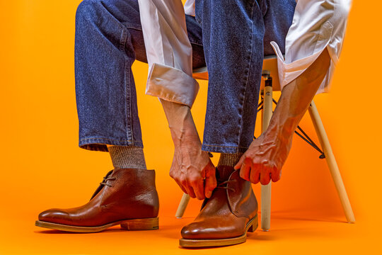Close-up of Man in Casual Jeans Clothing Wearing Leather Tanned Derby Boots Putting On Shoes Ad Tiying Up Laces Against Orange