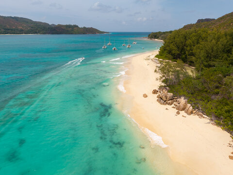 Aerial view of the beach where the turquoise waters meet the soft sand, boats bob gently, and lush green hills rise in the distance, Baie Ste Anne, Seychelles.