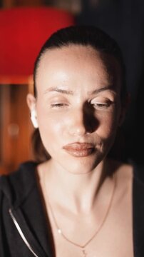 Cheerful brunette woman with a high ponytail in headphones applying contour and bronzer with a large brush on her face, natural look, smiling at night in her apartment with red lamp, vertical video