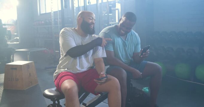 Two African men sitting at gym bench checking phones, showing funny clip, sharing laughter