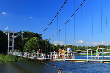 People and children walking and relaxing on a wire suspension bridge over the lake in a public park on a sunny day.