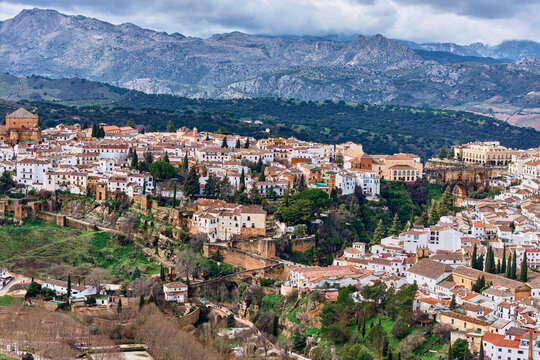 Aerial view of sun-kissed buildings cascading down the hillside, a tapestry of terra cotta roofs against the backdrop of the majestic mountains, Ronda, Andalusia, Spain.