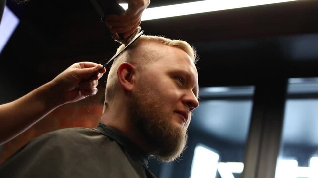 Barber cutting a man&rsquo;s hair with clippers in a barbershop. Side profile of a male client with short haircut and beard, professional grooming service in a salon.