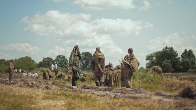 Wide shot shows uniformed reenactors in camouflage preparing for Great Patriotic War event. Soldiers stand and sit in open field, with trees and cloudy sky as backdrop. Summer day