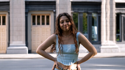 A portrait of a cheerful girl with long curly hair smiling at the camera. A close-up portrait of a friendly young woman in casual clothes against a blurred city background. Outdoors.