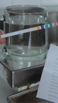 Close-up of human hand holding simple test strip submerged in glass of tap water for analysis of overall water hardness. Comparison chart for result determination in domestic setting. Vertical shot