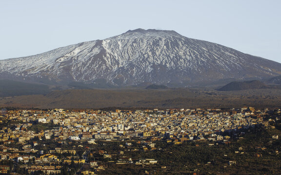 View of snow-dusted Mount Etna looms over the warm-toned cityscape with the houses nestled amidst the verdant foothills, Bronte, Sicily, Italy.