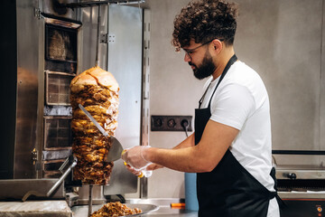 Male chef in black apron slices meat from vertical rotisserie in modern kitchen, stainless steel...