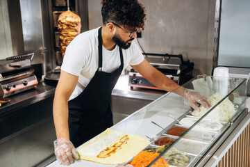 Male chef in black apron prepares shawarma wrap with fresh ingredients at a modern restaurant kitchen, grill and food containers visible in the background