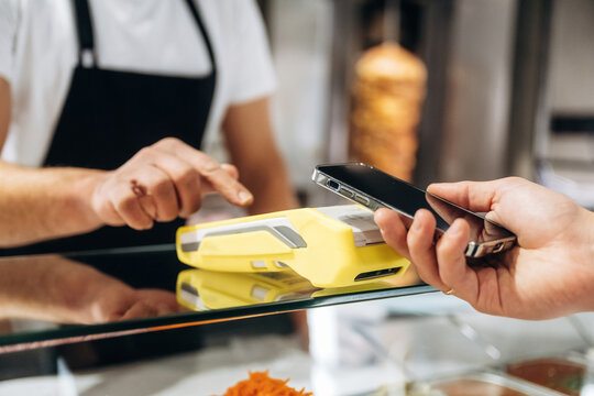 Customer using smartphone for contactless payment at food counter with cashier pointing towards payment terminal in a modern fast-food setting