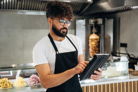 Male restaurant worker in black apron using tablet to take orders in a fast-casual dining establishment with food items displayed in the background