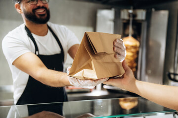 Male food service worker in black apron hands over a takeout bag to a customer at a counter in a restaurant with food preparation equipment visible in the background