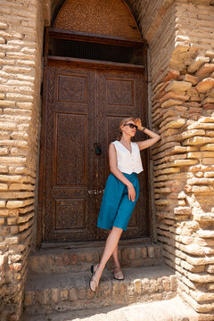 Young woman wearing sunglasses and casual clothes stands near an ancient carved wooden door of a brick building during, sunny day in a historical city while leaning against textured stone wall