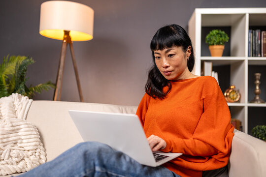 Asian Woman Working on Laptop at Home, Lifestyle and Freelance Concept. Middle Aged Female Sitting on Sofa Using Computer for Remote Work or Browsing the Internet in Living Room Apartment.