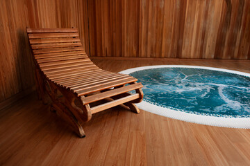Wooden lounge chair stands on a timber floor next to a bubbling round hydromassage pool with blue water in a modern indoor spa area featuring, natural wood wall panels and warm ambient lighting © timofeev
