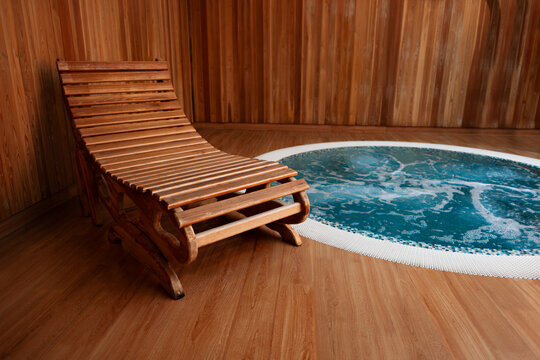 Wooden lounge chair stands on a timber floor next to a bubbling round hydromassage pool with blue water in a modern indoor spa area featuring, natural wood wall panels and warm ambient lighting