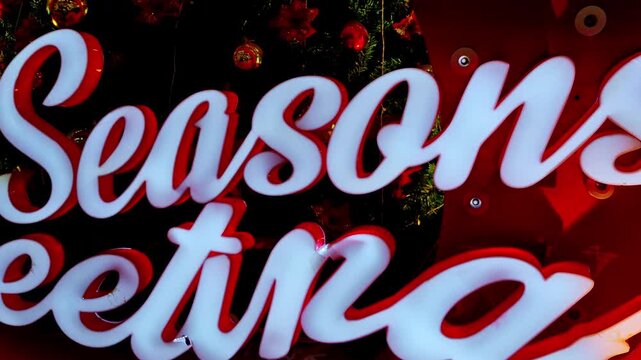 A glowing white and red Season's Greetings sign is displayed in front of a decorated Christmas tree. The bright, cursive lettering stands out against the dark greenery and festive ornaments.