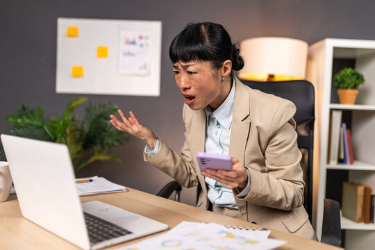 Frustrated Asian businesswoman shouting at laptop. Angry employee with smartphone, Workplace Stress concept. Stressed woman in beige blazer gesturing with hand while looking at computer screen.