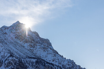 Sunlight bursts from behind the jagged, snow-dusted ridges of the Pomagagnon range north of Cortina d Ampezzo, creating a brilliant lens flare against a pale, hazy winter sky over the Dolomites.