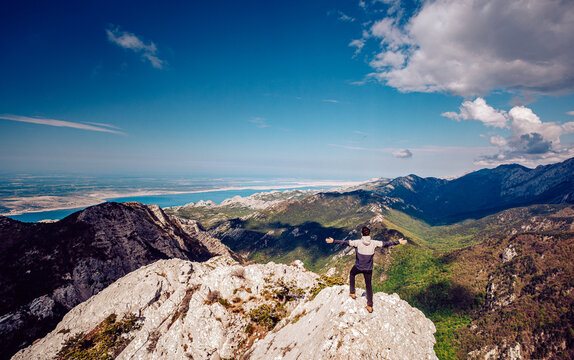 View of a lone figure standing triumphant on a rocky peak, arms outstretched against the backdrop of distant mountains and a serene coastline, Krasno, Li&Auml;ko-senjska &Aring;&frac34;upanija, Croatia.