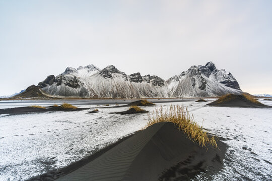 View of stark black sand dunes meet icy white snow under towering, rugged mountains creating a mesmerizing contrast, Hofn, Iceland.