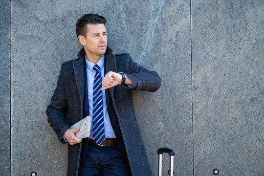 Businessman with suitcase standing at a wall checking the time