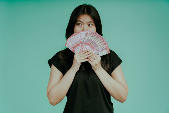 Big Profit. Happy young chinese woman covering face with rupiah cash fan. Lucky millennial korean female holding a lots of money, gor prize, standing isolated on green background, Closeup shot.