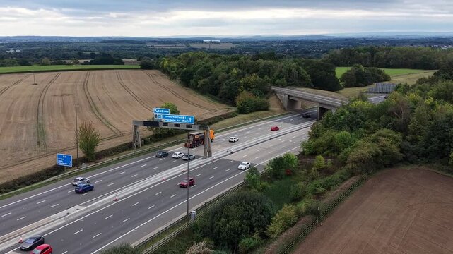 Aerial drone pull out video showing a motorway near Banbury in Oxfordshire, England, with moving cars, road signs, surrounding fields, trees, and countryside landscape.