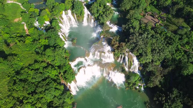 Breath-taking drone shot of the multi-level Ban Gioc Waterfall, featuring a visible rainbow over the turquoise water and small boats navigating the river.