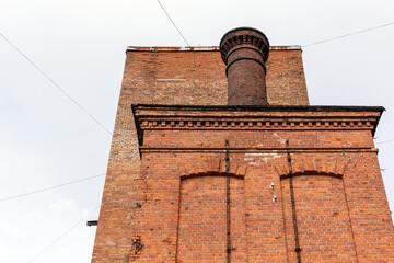 Prominent red brick chimney rises above a historic factory facade, showcasing industrial architecture, weathered masonry and urban heritage details under a pale sky