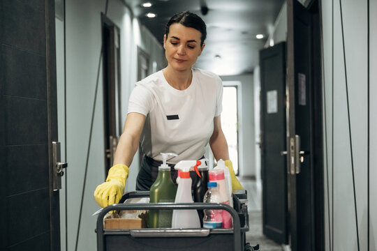 Female hotel housekeeper in white shirt and yellow gloves pushes cleaning cart filled with supplies down a modern hotel corridor with doors and lighting visible