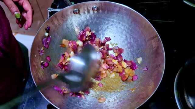 A top-down action shot of chopped red onions being saut�ed in a hammered metal wok. A large spoon is caught in motion, stirring the onions as they soften and brown over a stove.