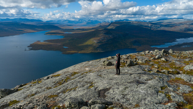 Aerial view of a lone figure stands atop a rocky peak overlooking the vast, still waters of a lake surrounded by rugged mountains in the distance, Kilpisj&Atilde;&curren;rvi, Lapland, Finland.
