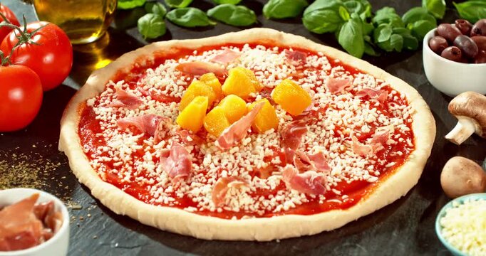 Super slow motion close-up of fresh pineapple pieces falling onto prepared Hawaiian pizza. Raw dough with tomato sauce, mozzarella and ham surrounded by ingredients. Concept of pizza preparation.