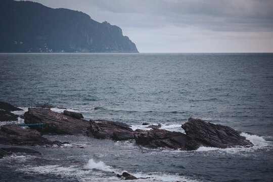 Waves crash against dark, jagged rocks as a stormy sky looms overhead. The coastline stretches into the distance, revealing a misty mountain range nearby. Nature's power is captivating