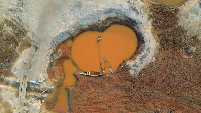 Aerial view of boats resting on bright orange water surrounded by cracked, dry earth in stark contrast to the sandy banks, Sunamganj, Sylhet Division, Bangladesh.
