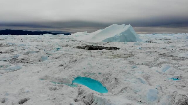Aerial view of pristine white icebergs scattered across the ocean, amidst grey skies contrasting with small turquoise pools, Greenland.