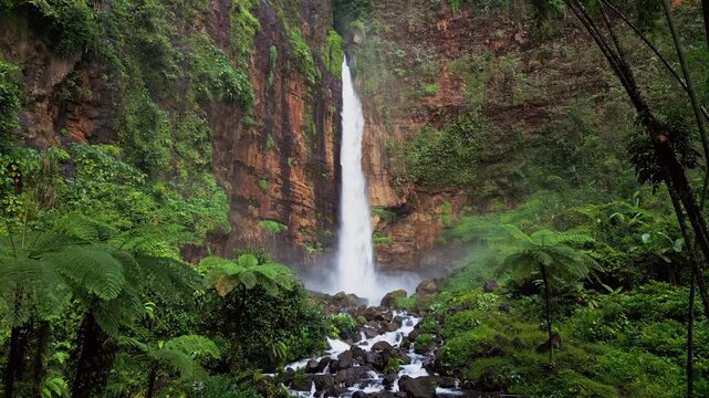 Drone footage shows Kapas Biru Waterfall in East Java dropping into a rocky river channel. Dense jungle surrounds the canyon as white water flows through dark stone, creating a dramatic tropical scene