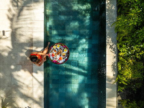 Aerial view of a woman lounging with a floating breakfast tray in the pool, framed by the geometric tiles and lush greenery, Prince Maurice, Flacq District, Mauritius.