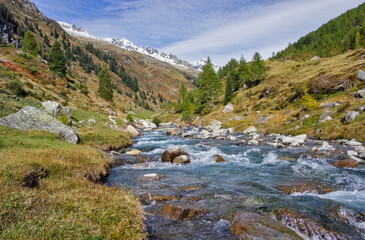 A picturesque mountain stream flows through a lush green valley in the South Tyrolean Alps, surrounded by autumn slopes and snow-capped peaks.