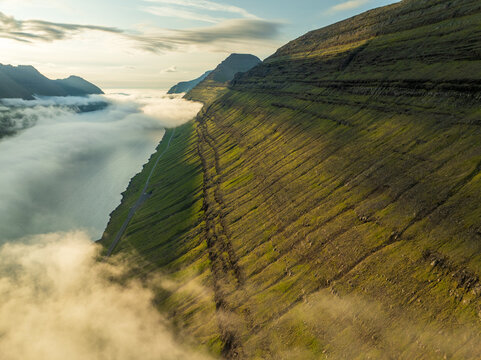 Aerial view of verdant slopes meeting the crisp, white fog snaking through the fjords, a dance of nature's elements, Hvannasund, Northern Isles, Faroe Islands.
