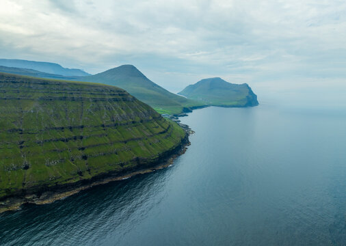 Aerial view of rugged cliffs meet the tranquil sea, emerald slopes contrasting with the deep blue waters, creating a serene coastal vista, Vidoy, Faroe Islands.