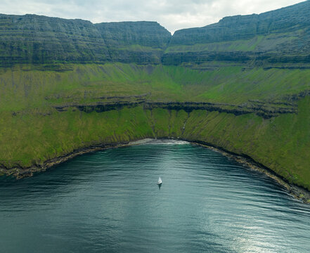 Aerial view of a lone sailboat glides on the water, nestled between towering cliffs of green and grey, a serene contrast of nature's elements, Vidoy, Faroe Islands.