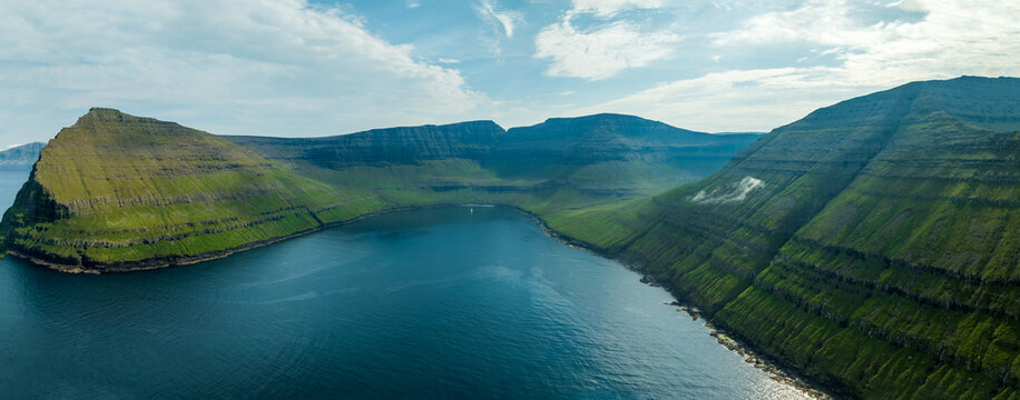 Aerial view of the dramatic coastline where the deep blue sea meets the lush green cliffs under a sky with scattered clouds, Vidoy, Northern Isles, Faroe Islands.