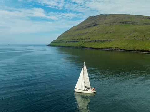 Aerial view of a sailboat gliding on the tranquil waters near the rugged coastline of Vidoy, Northern Isles, Faroe Islands.