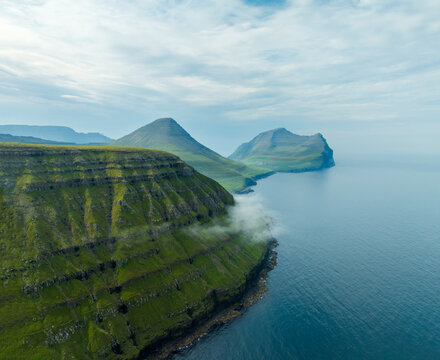 Aerial view of dramatic cliffs meet the tranquil sea, shrouded in mist, a serene dance of earth and water unfolds, Vidoy, Faroe Islands.