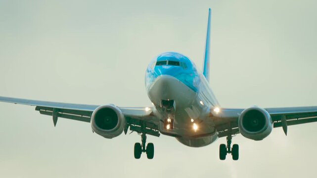 Jet flying low with landing gear extended and engines visible against cloudy background. Airplane approaching runway with blue nose and tail through overcast sky. Passenger aircraft descending in