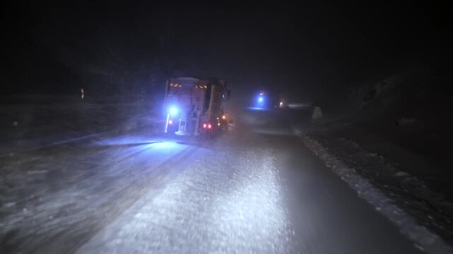 Two snowplow trucks clearing deep snow on a highway at night in dense fog and heavy snowfall. Blue and amber lights illuminate drifting snow, capturing extreme winter weather and road maintenance oper
